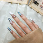 Short press on nails on hand: nude pink nails with blue glitter, white flower details, gold rings