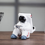 Astronaut resin phone stand figurine holding a rose-gold smartphone on a desk