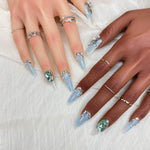 Long stiletto press-on nails with blue ombre tips, silver details, and emerald crystal