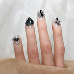Short press-on nails with dark teardrop gems, silver glitter, and bow charms