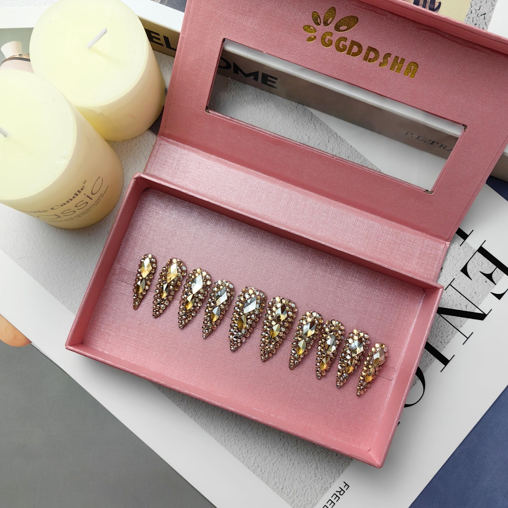 Almond press-on nails arranged in a pink display box, gold and clear rhinestone accents