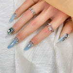Square press-on nails with light blue tips, silver accents, and green oval crystal